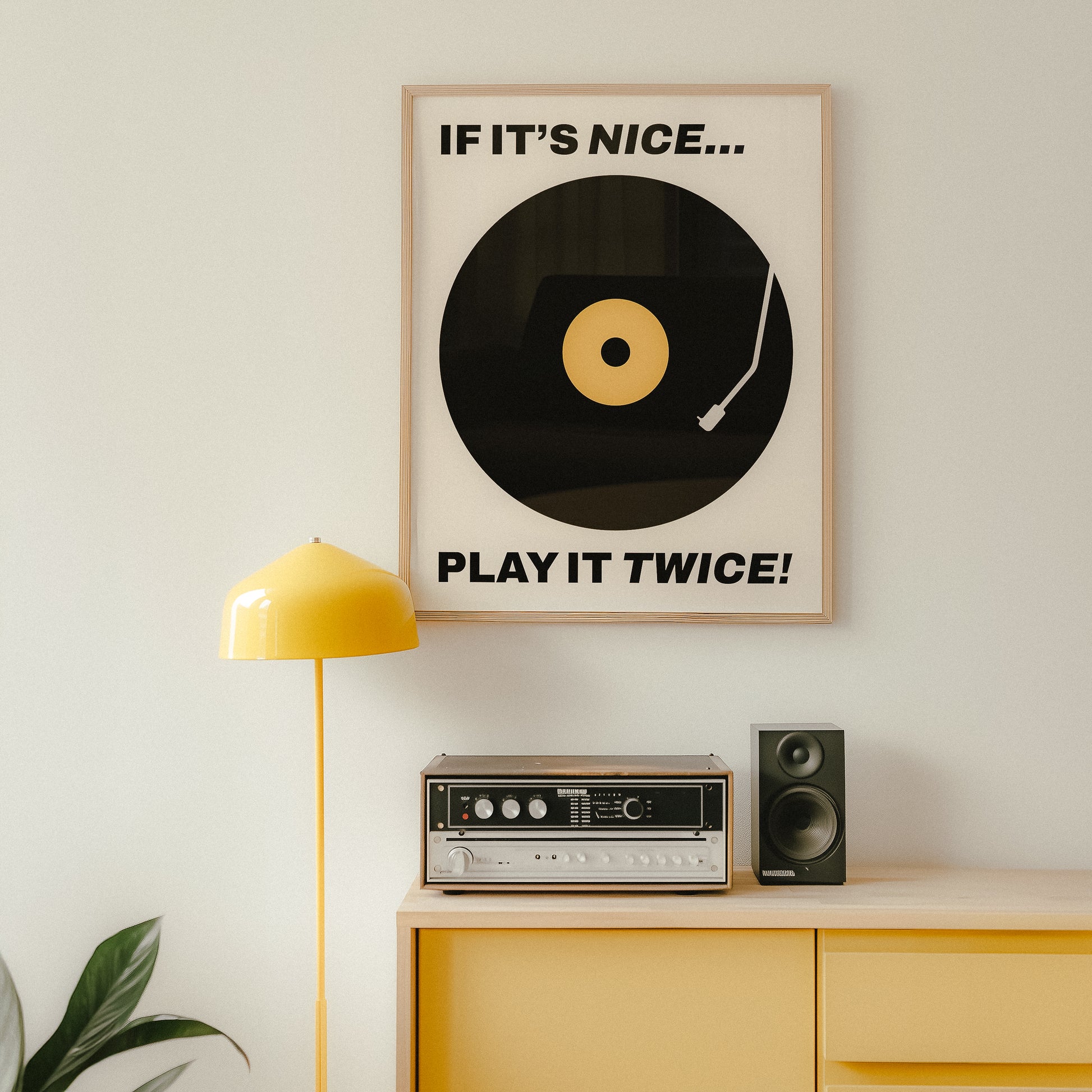 Framed typography poster featuring a bold black vinyl record design with a yellow label, accompanied by the phrase 'IF IT'S NICE... PLAY IT TWICE!' in striking black font. Midcentury modern themed wall art.